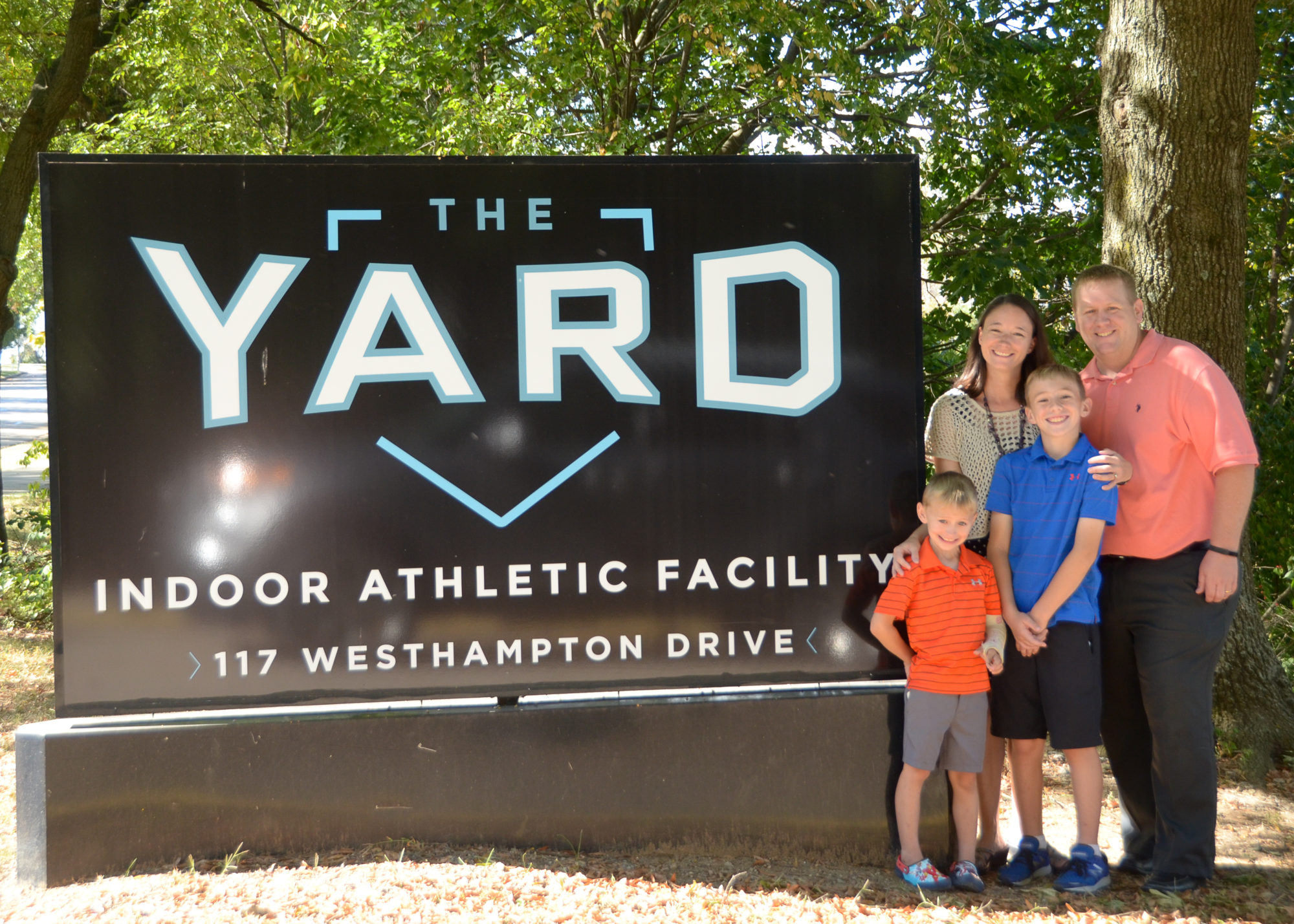 Nick Williams and his family standing in front of the roadside sign that reads "The Yard Indoor Athletic Facility, 117 Westhampton Drive" with trees in the background.