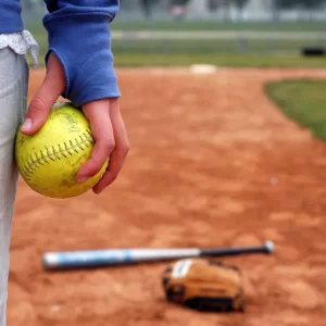 Child holding a softball on a dirt field with a bat and glove in the background.